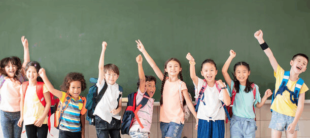 Gruppe von Grundschulkindern verschiedener Herkunft steht lachend vor einer Schultafel und feiert den Schulstart.