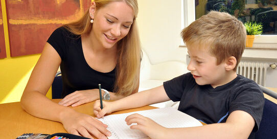Junge lernt mit Nachhilfelehrerin am Tisch. Gemeinsam bearbeiten sie Aufgaben in einem Heft.